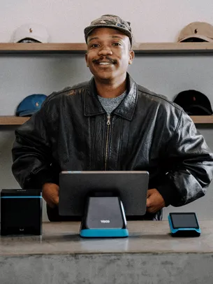 A smiling merchant at a cap and accessories store, with a complete Yoco POS system set up on the counter in front of them.