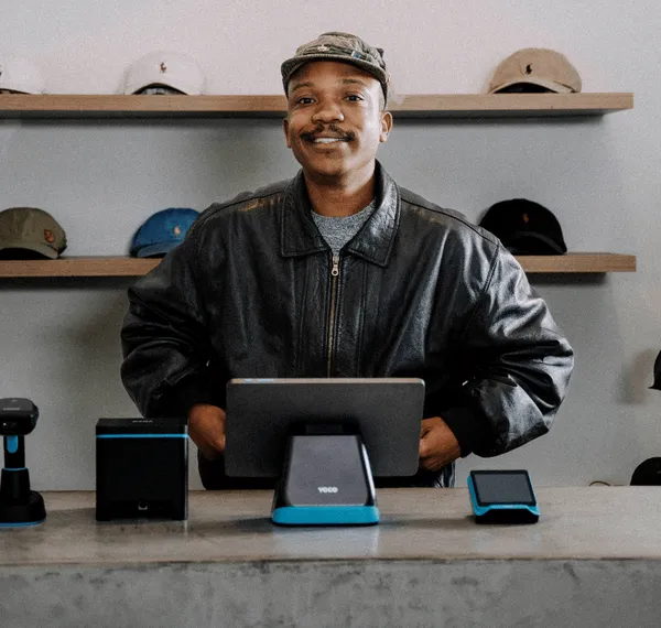 A smiling merchant at a cap and accessories store, with a complete Yoco POS system set up on the counter in front of them.