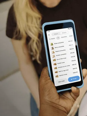 Handheld POS device showing a product menu and prices, held by a merchant while presenting items to a seated customer, illustrating mobile order taking and checkout.