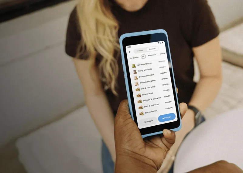 Handheld POS device showing a product menu and prices, held by a merchant while presenting items to a seated customer, illustrating mobile order taking and checkout.