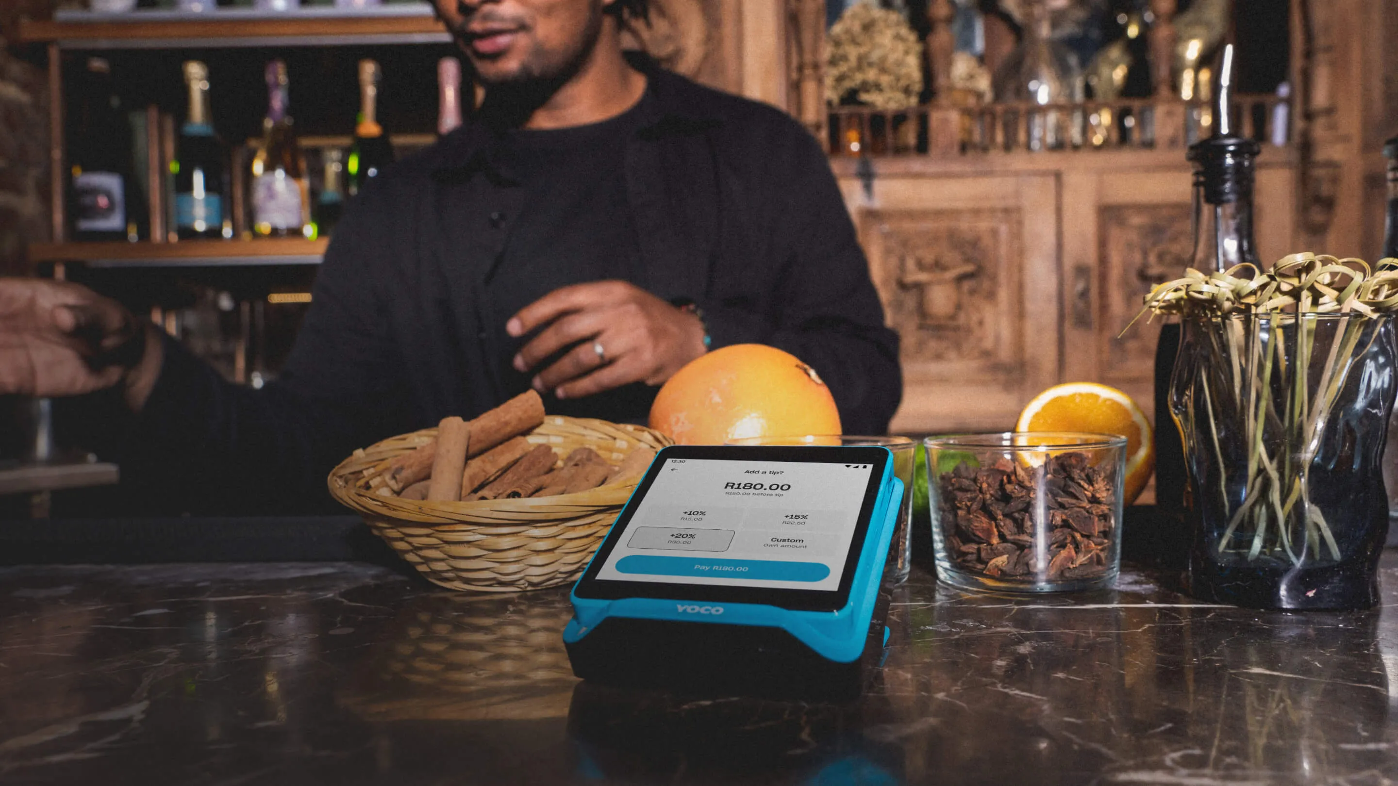 Yoco Khumo Print card machine on a bar counter, displaying a payment screen, with spices and a bartender in the background.