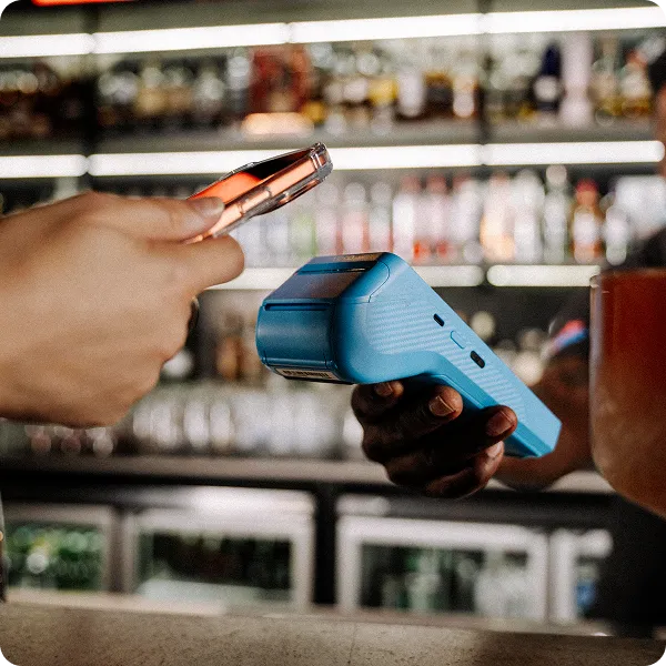 Handheld POS terminal accepting a contactless payment, with a customer tapping a smartphone against a Yoco card reader at a bar counter.