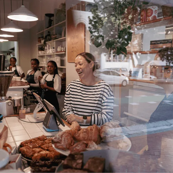 Smiling café owner behind a pastry counter with staff working in the background.