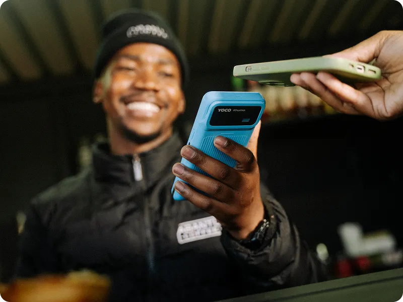 Merchant holding a handheld POS terminal while a customer makes a tap-to-pay transaction with a smartphone.
