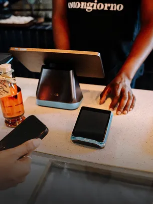 Retail checkout scene with a handheld POS device on the counter, as a customer prepares to pay with a smartphone while a cashier stands behind a register, illustrating in-store digital payments.