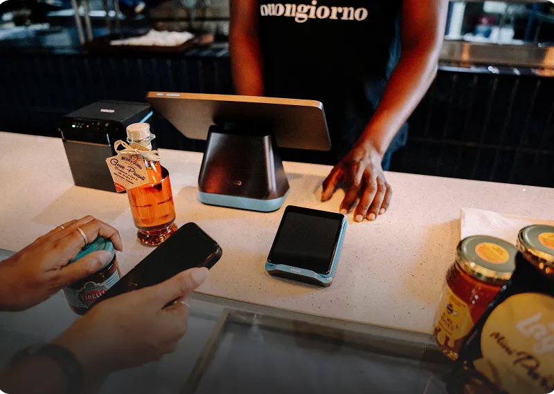 Retail checkout scene with a handheld POS device on the counter, as a customer prepares to pay with a smartphone while a cashier stands behind a register, illustrating in-store digital payments.