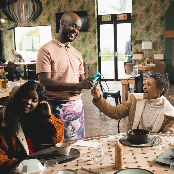 A waiter using a Yoco POS device to assist two customers making a payment at a café.
